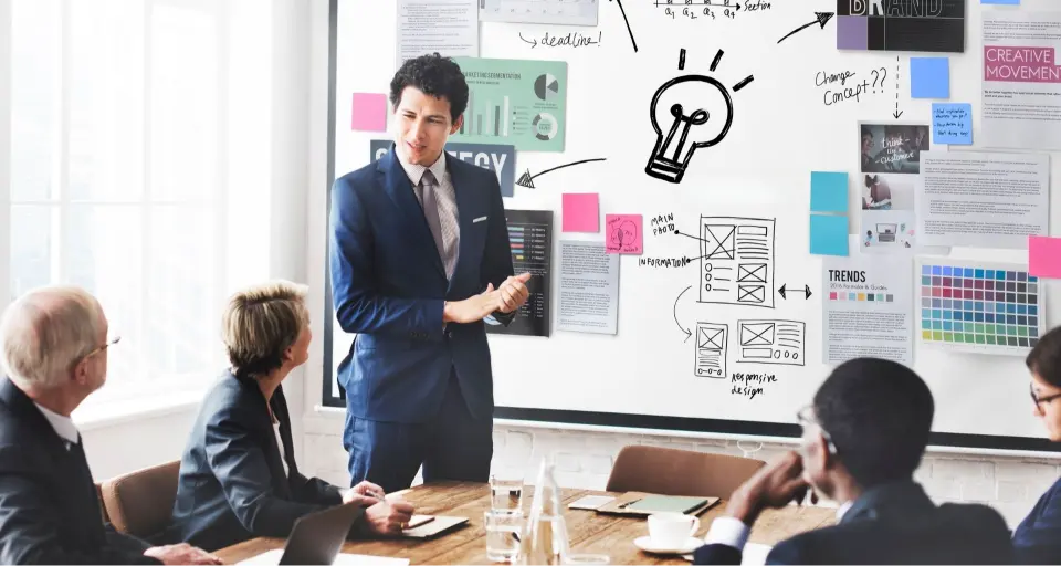 Professionals discuss strategy; man stands by whiteboard with diagrams and light bulb sketch.