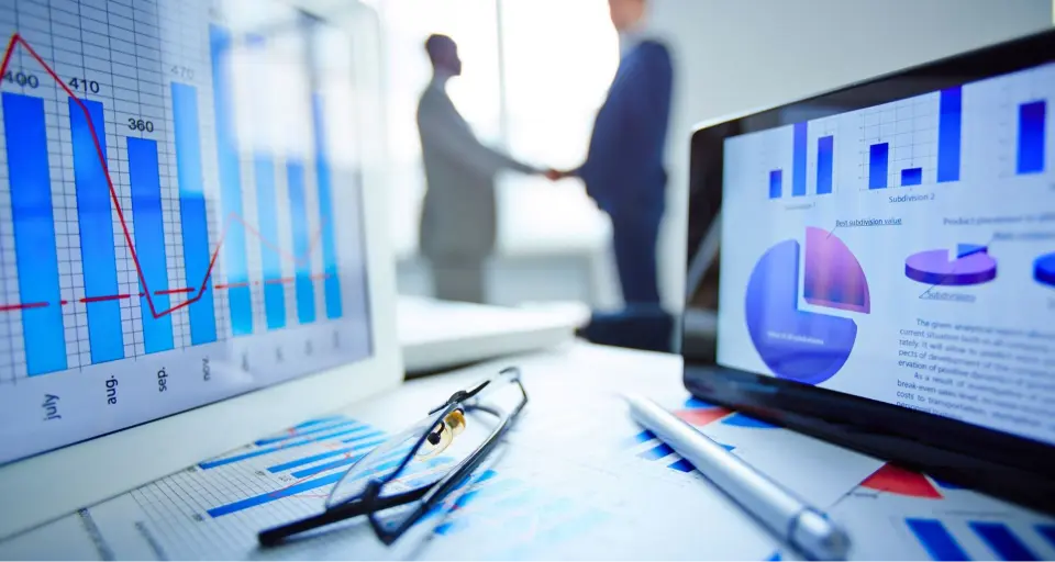 Close-up of financial charts, glasses, and a pen, with two people shaking hands in the background.