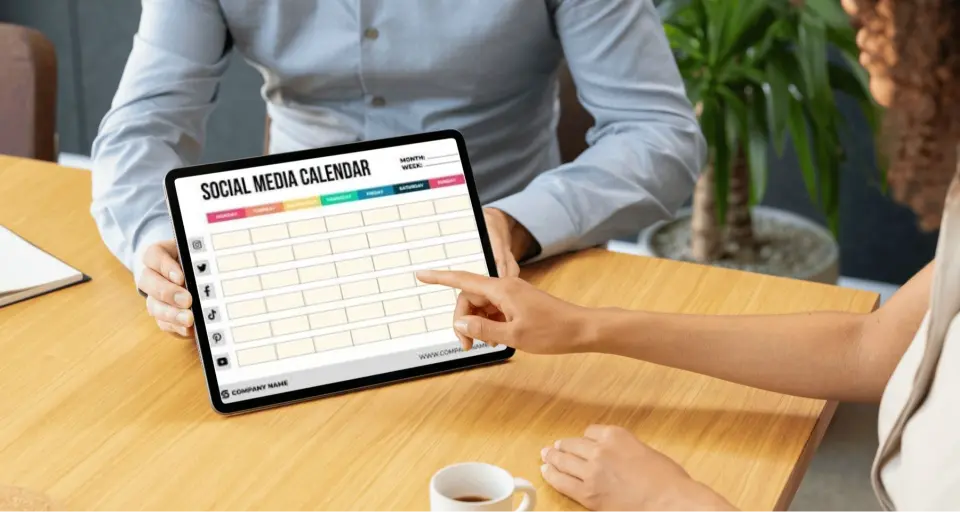 Two people review a social media calendar on a tablet, one pointing, with coffee on the table.