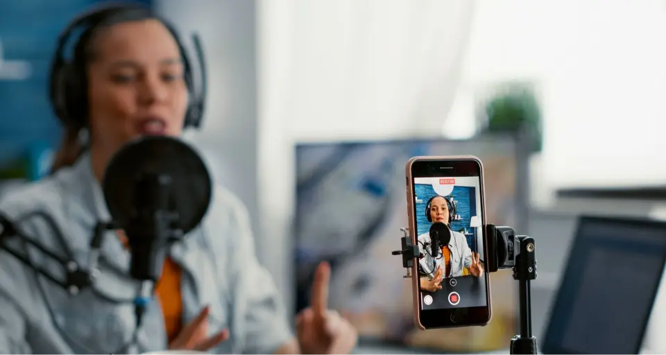 Person speaks into a microphone with headphones, recorded on a smartphone mounted on a tripod.